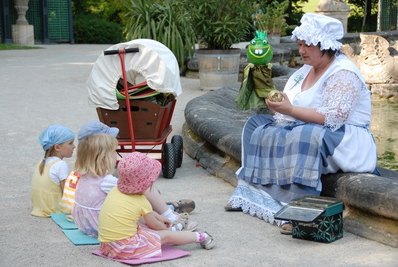 Eine Frau sitzt am Brunnenrand mit einer Froschpuppe in der Hand, davor sitzen 4 Kinder