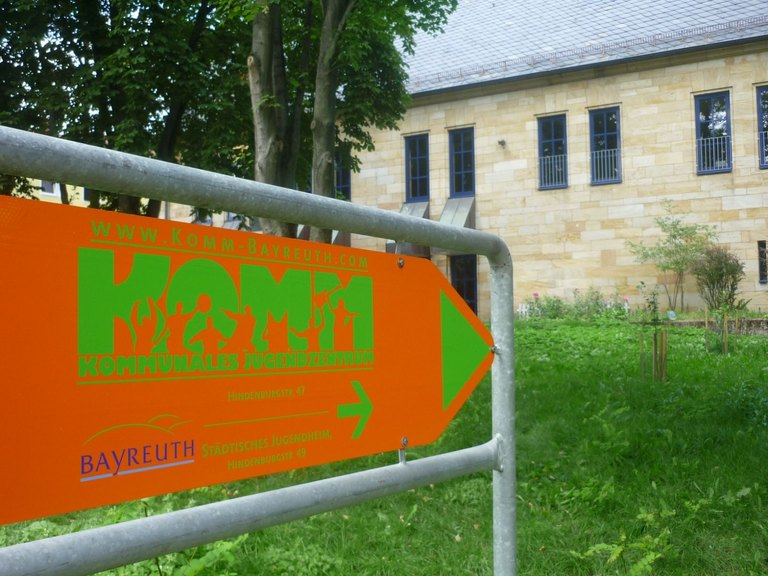 Hinweisschild des Kommunalen Jugendzentrums mit grünem Logo auf orangem Grund vor dem Gebäude