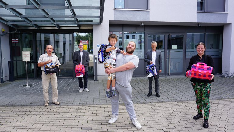 Vorstellung der Schultaschen von vier Maennern und einer Frau, ein Junge sitzt auf dem Arm seines Vaters