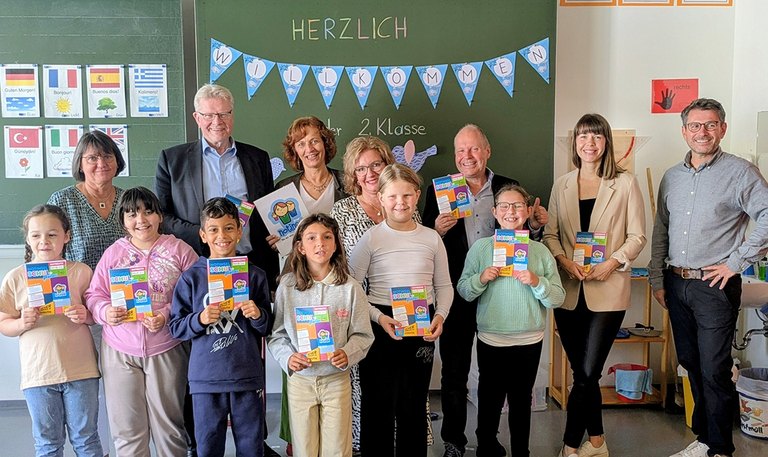 Eine Gruppe von Erwachsenen und Kindern steht in einem Klassenzimmer vor einer grünen Tafel und präsentiert das neue Notinselhausaufgabenheft. 