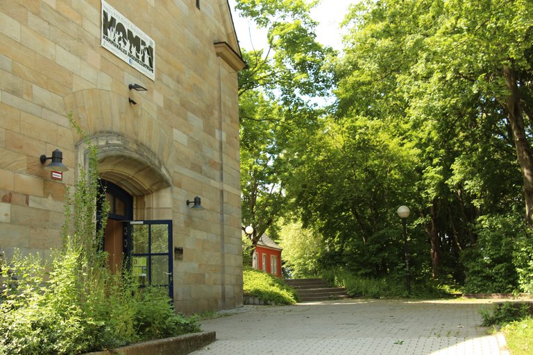 Außenfassade des Kommunalen Jugendzentrums Bayreuth mit grünen Bäumen und Sträuchern vor der Eingangstüre