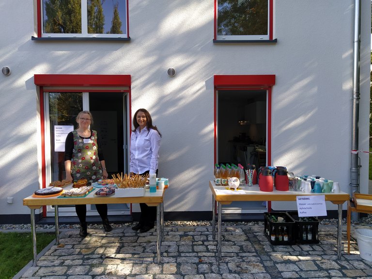 Zwei Frauen, die hinter einem Tisch mit Kuchen vor einem Haus stehen