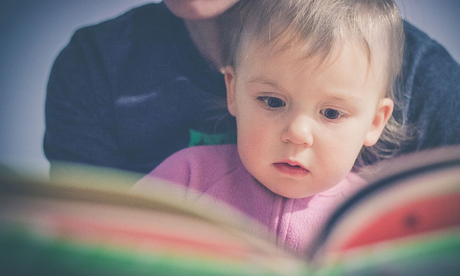 ein aufgeschlagenes Buch mit einem Kleinkind davor, dahinter eine Person, die vorliest